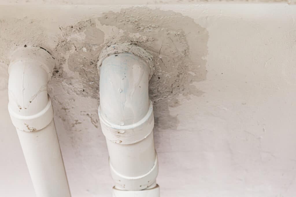 Zwei weiße Kunststoffrohre, die durch eine Decke führen, mit sichtbaren Ausbesserungen und Rissen im Putz um die Durchführungen