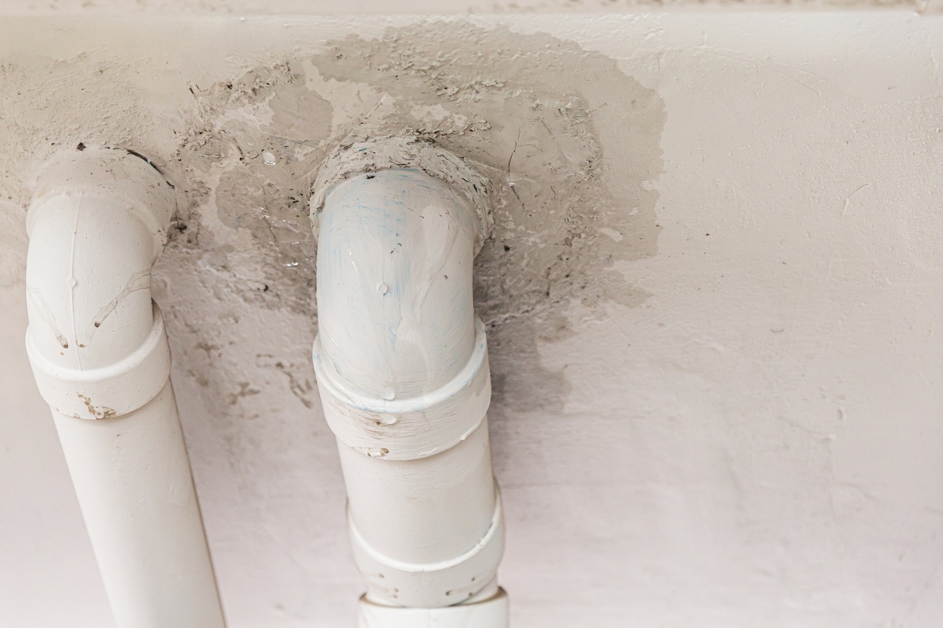 Zwei weiße Kunststoffrohre, die durch eine Decke führen, mit sichtbaren Ausbesserungen und Rissen im Putz um die Durchführungen