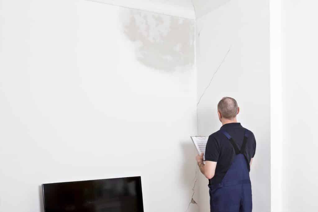 Handwerker in Arbeitskleidung steht vor einer Wand mit großem Riss und hält ein Klemmbrett